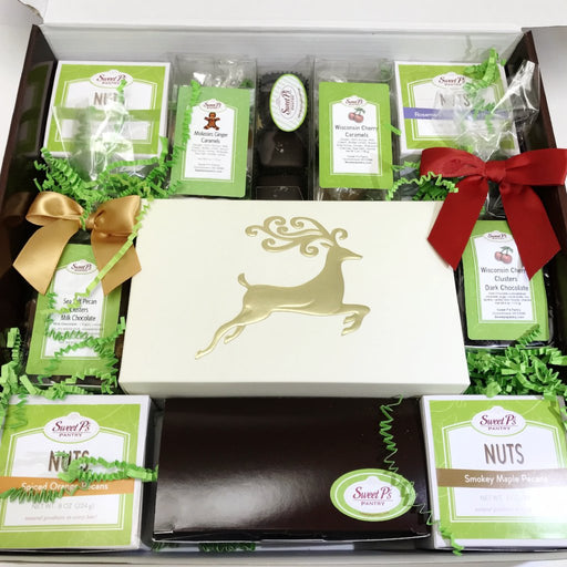 A gift assortment box containing various packaged snacks and treats, including cashew almond toffee, candied pecans, sea salt caramels, cherry caramels, seasonal caramels, toffee, nut mix, and chocolate pecan clusters, adorned with a gold-colored deer logo on the lid and green and red decorative ribbons.